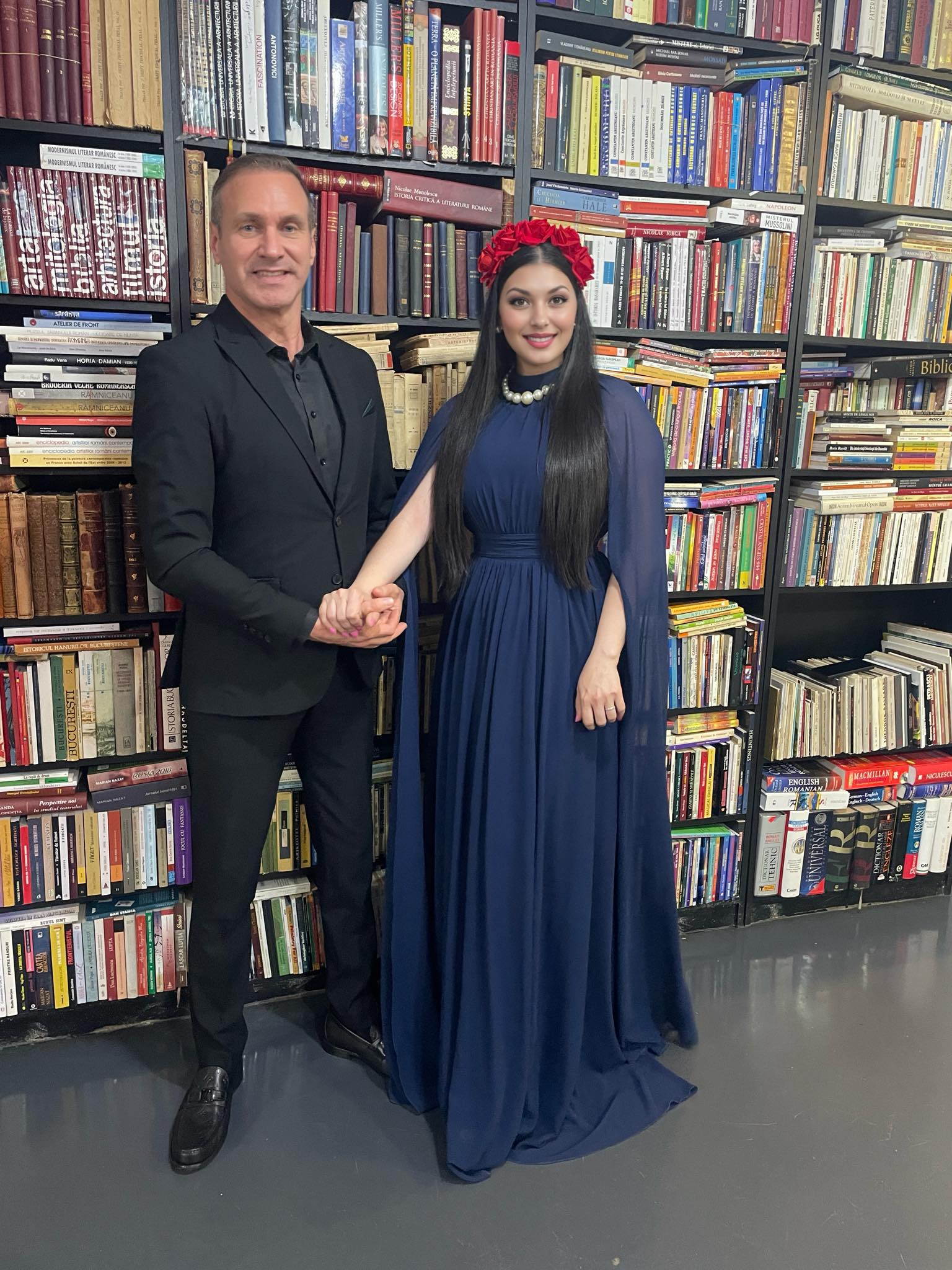 Alin Oprea în costum negru și Medana în rochie lungă, mov, cu o coroniță de flori pe cap, ținându-se de mână în fața unui raft plin cu cărți