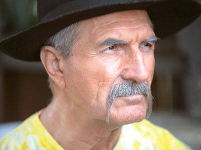 Gheorghe Dinică în rolul lui Aurică Fieraru din serialul „Inimă de Țigan”, cu mustață lungă, pălărie pe cap și cămașă galbenă