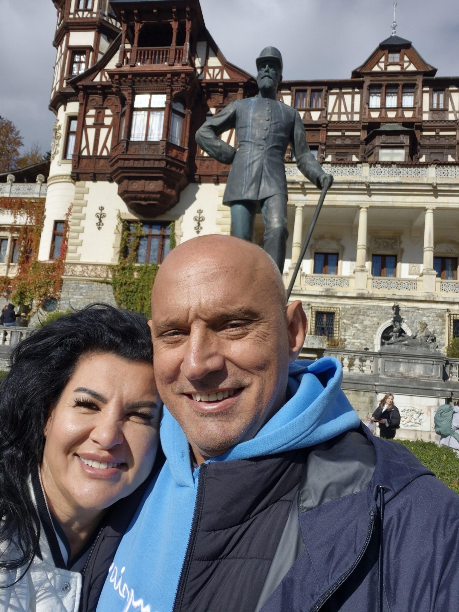 Florin Prunea și Lăcrămioara Ursu, zâmbitori, îmbrăcați în haine lejere, făcându-și un selfie împreună în fața Castelului Peleș