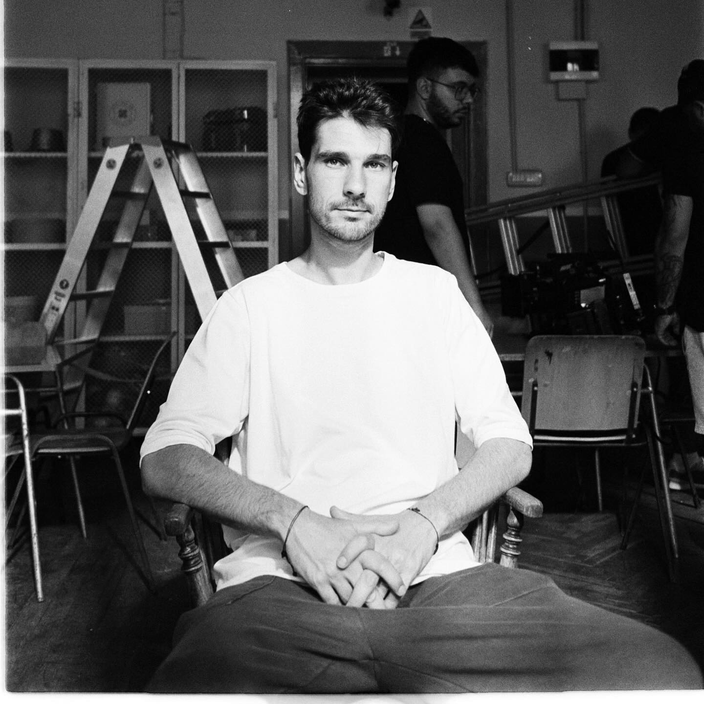 Imagine alb-negru cu Anghel Damian serios, stând pe un scaun, în timp ce în spatele său colegii cară recuzita