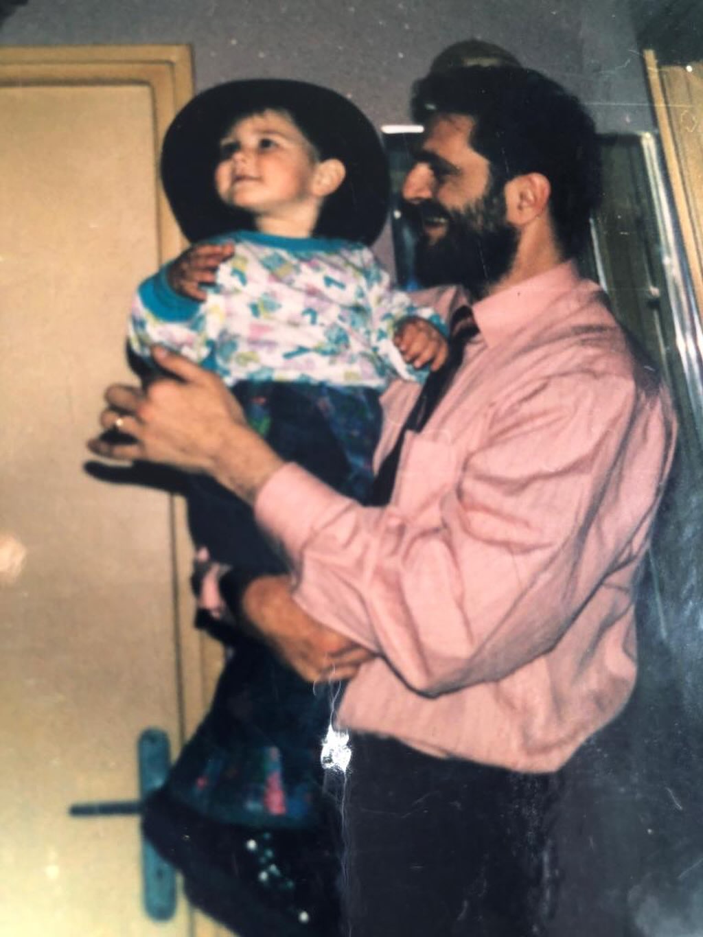 Anghel Damian în copilărie, îmbrăcat în blugi și bluză colorată, cu pălărie pe cap, în brațele tatălui său, Laurențiu Damian