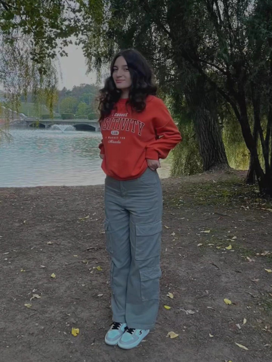 Alexandra Neagu în parc, pe malul unui lac, îmbrăcată în pantaloni gri și bluză roșie