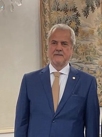 Adrian Năstase serios, stând drept la poză, lângă un tablou de mari dimensiuni, îmbrăcat într-un costum bleumarin, pe sub care a pus o cămașă bleu și pe care l-a accesorizat cu o cravată nude