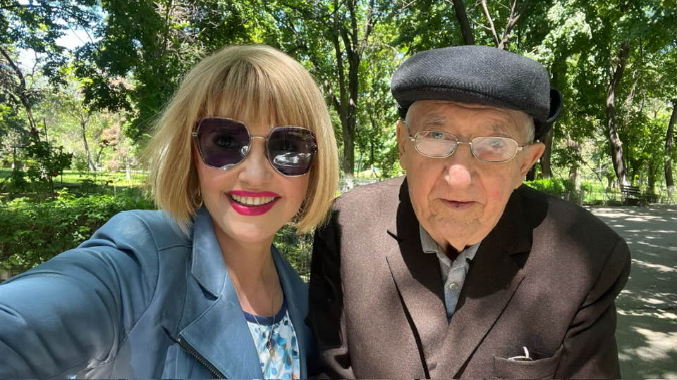 Marina Almășan cu ochelarii de soare la ochi, în parc, alături de tatăl ei, pe care l-a scos la plimbare, în costum închis la culoare, cu beretă pe cap