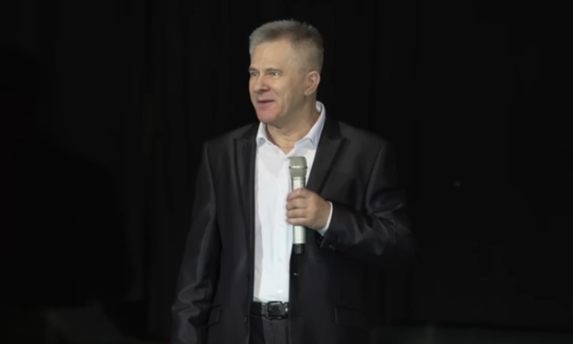 Doru Octavian Dumitru pe scenă, cu microfonul în mână, îmbrăcat în costum negru și cămașă albă