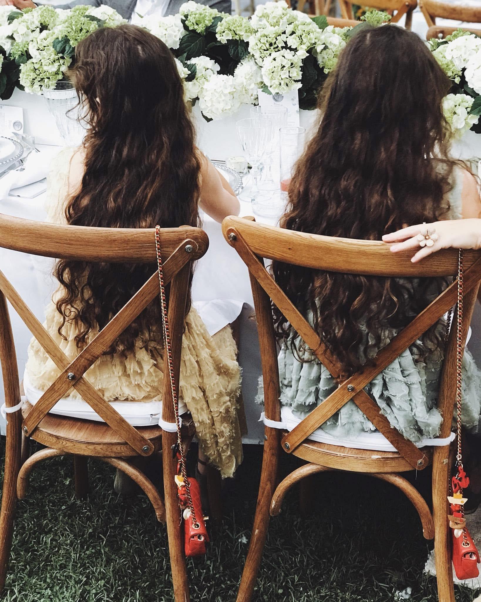 Gemenele Danei Budeanu fotografiate de la spate, îmbrăcate în rochii vaporoase în culori pastelate, stând la o masă lungă decorată cu flori albe