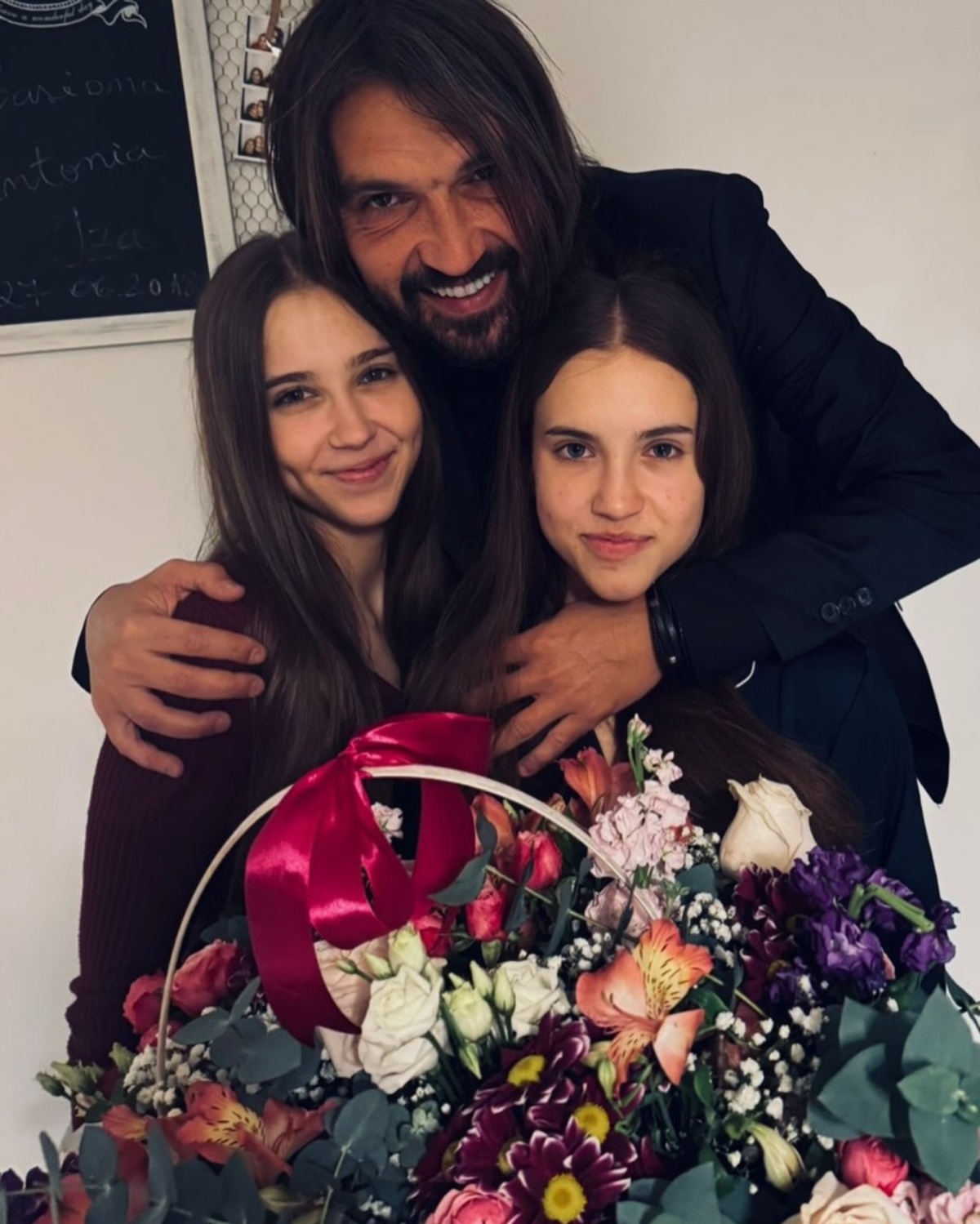 Dan Alexa zâmbitor, alături de fiicele sale, pe care le ține în brațe, în timp ce ele țin în poală un coș imens cu flori