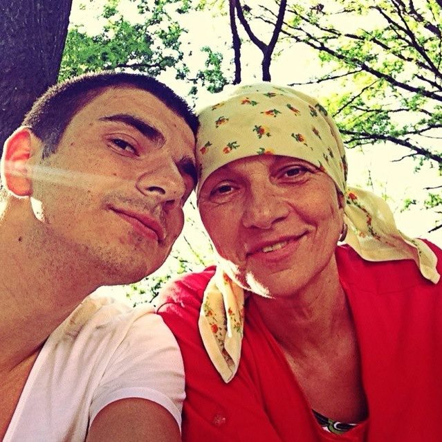 Alex Bogdan tuns scurt, în tricou alb, stând cu capul lipit de mama lui, îmbrăcată în roșu, cu batic pe cap, ascunzând urmele lăsate de chimioterapie