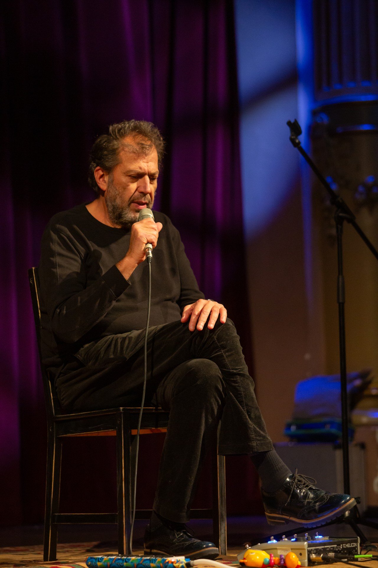 Artan pe scenă, în timpul unui concert, stând pe scaun, picior peste picior, cu o mână pe microfon și una pe genunchi