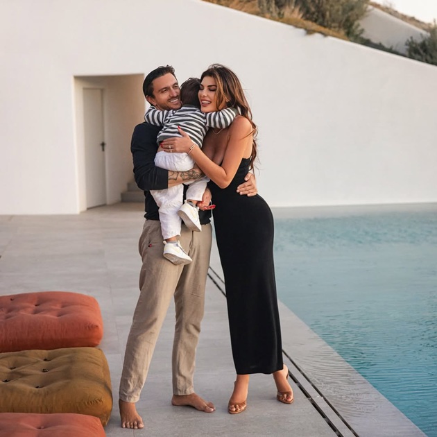  Angela Martini și Dragoș Săvulescu zâmbitori, îmbrăcați asortat, ținându-și fiul în brațe, pe marginea unei piscine