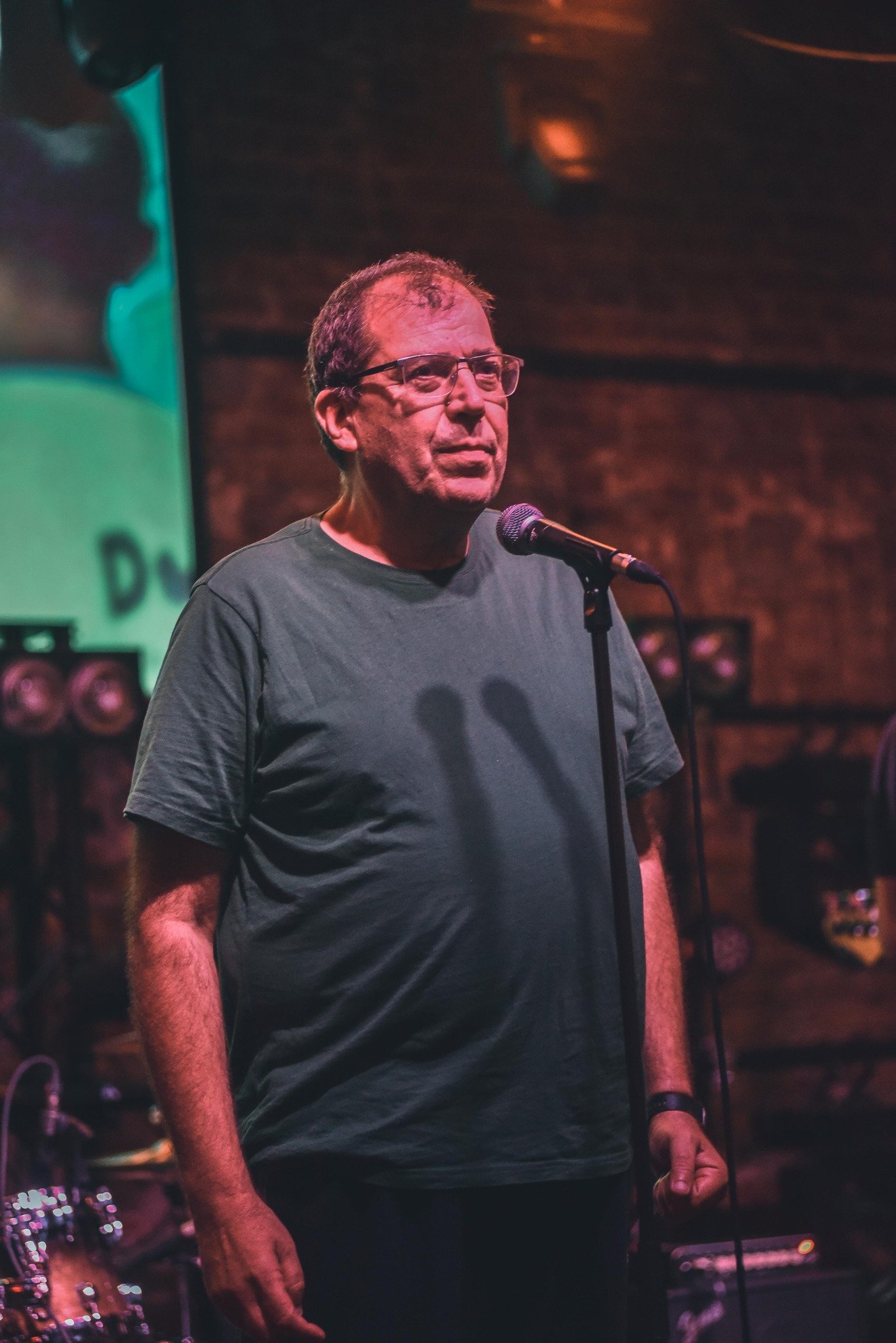 Artan pe scenă, într-un tricou gri, cu ochelarii de vedere la ochi, în fața publicului său, cu microfonul lângă el, pregătit să îi încânte pe cei din sală