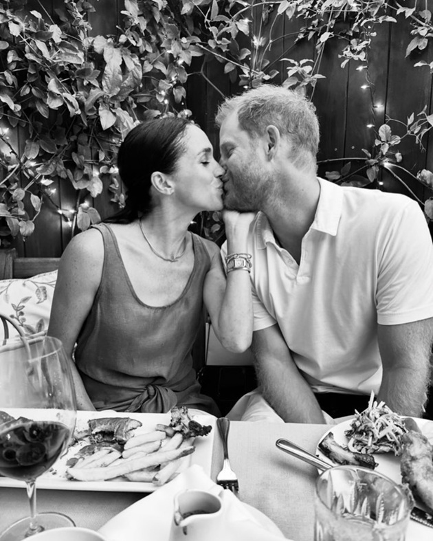 Meghan Markle și Prințul Harry sărutându-se în timp ce stau împreună la masa unui restaurant, ai cărui pereți sunt decorați cu plante cățărătoare și lumini