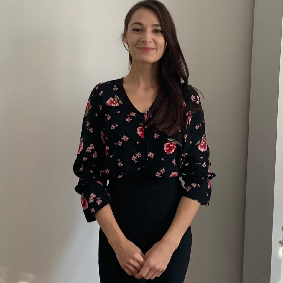 Monica Cioata zâmbitoare, îmbrăcată în pantaloni negri și bluză neagră cu flori, ținându-și mâinile împreunate și părul pe o parte