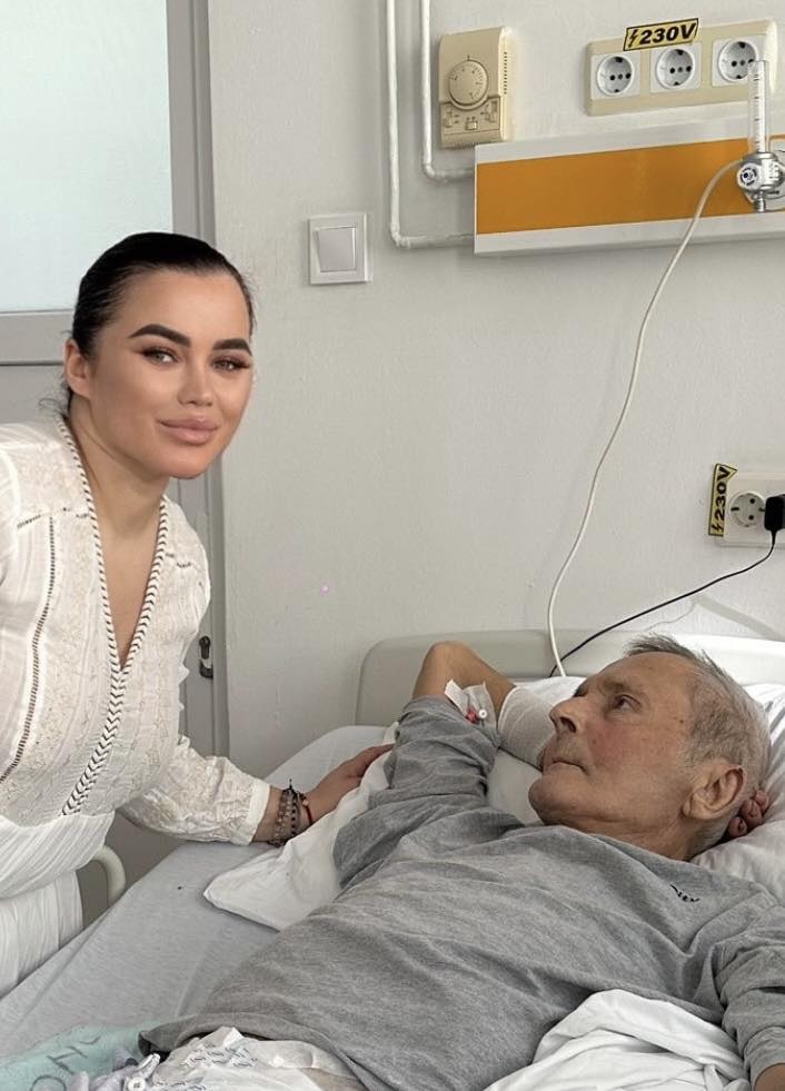 Carmen de la Sălciua îmbrăcată în alb, cu părul prins la spate, stând alături de tatăl ei aflat pe patul de spital, îmbrăcat în pijamale gri, cu branula în mână