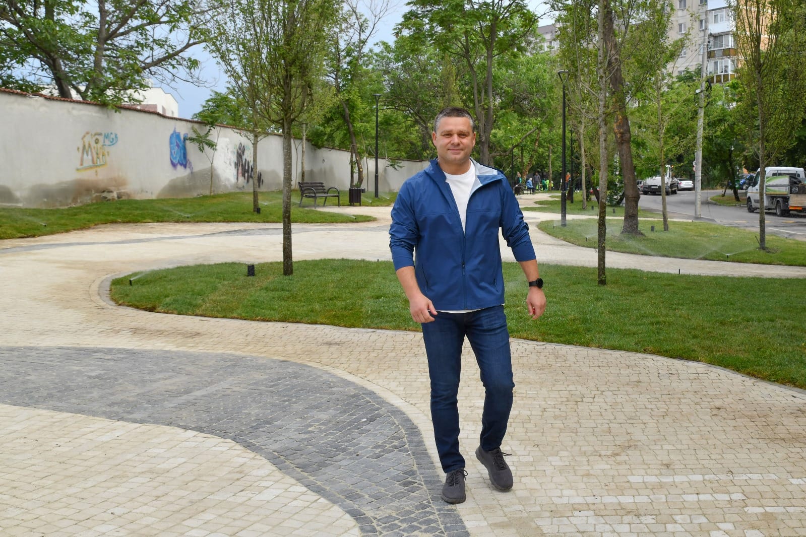 Ciprian Ciucu plimbându-se zâmbitor într-un spațiu verde din sectorul 6, pe aleea unui parc cu multă verdeață și mulți copaci, îmbrăcat în alb și mai multe nuanțe de albastru