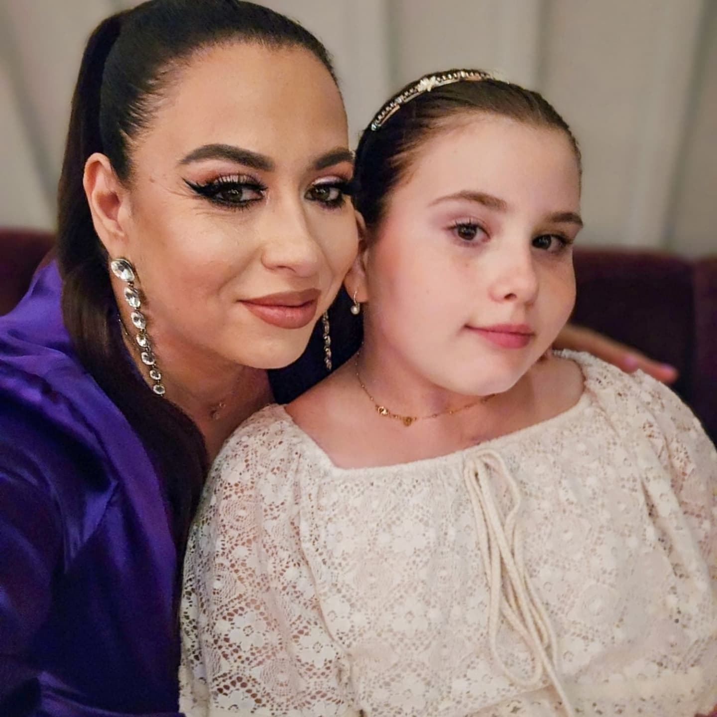 Oana Roman îmbrăcată în mov, purtând cercei lungi cu pietre, stând în spatele fiicei sale, cu o mână pe umărul ei, în timp ce Isabela este îmbrăcată într-o rochie albă din dantelă