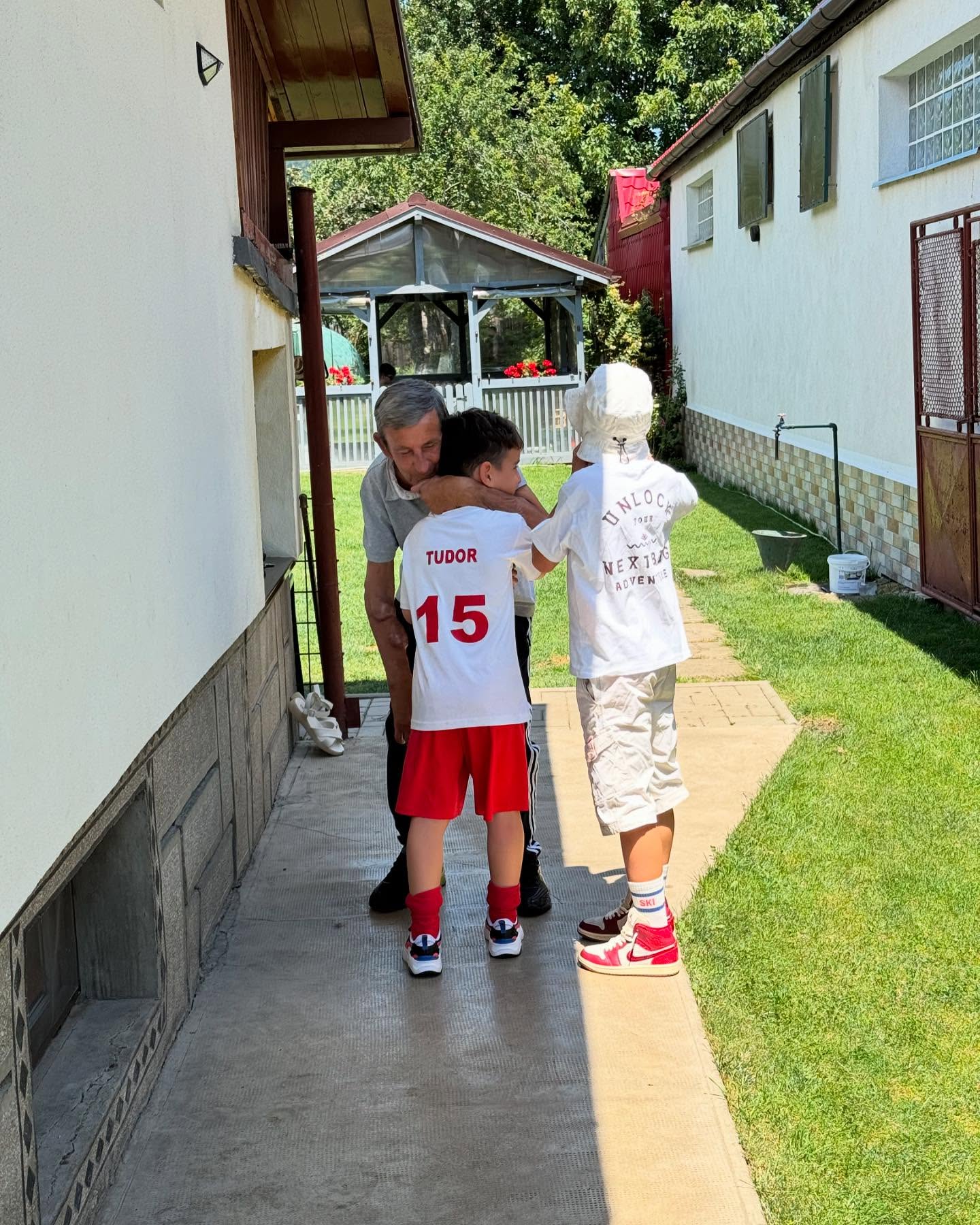 Tatăl Ioanei Blaj îmbrățișându-i pe copiii ei, Tudor și Filip, în curte, la umbră, într-o zi însorită de vară