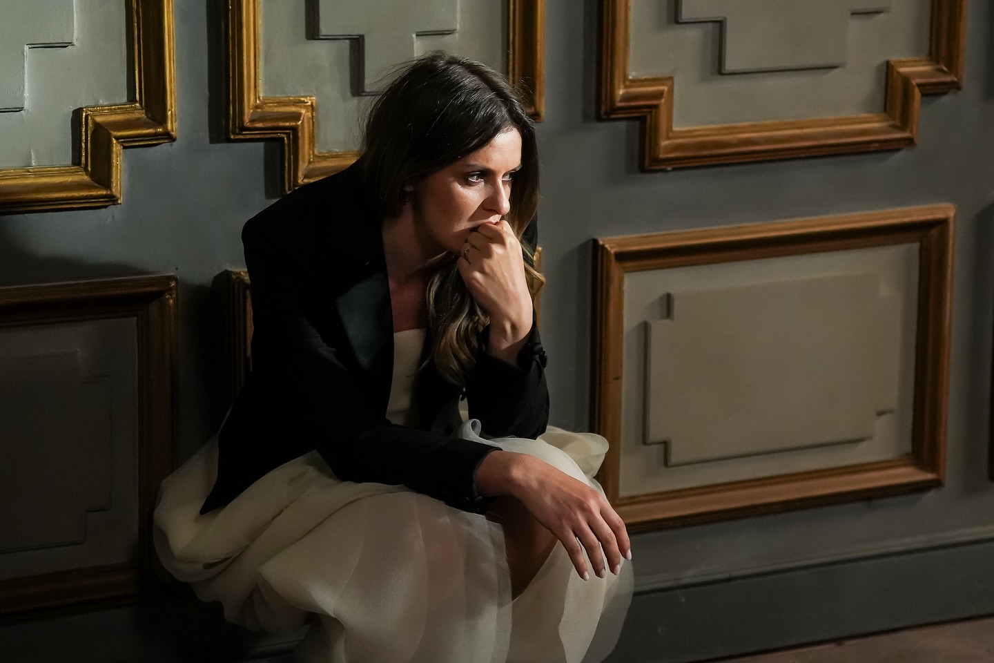 Ioana Blaj în personajul Alice din „Lia, soția soțului meu”, tristă și îngândurată, stând sprijinită de un perete gri cu model auriu