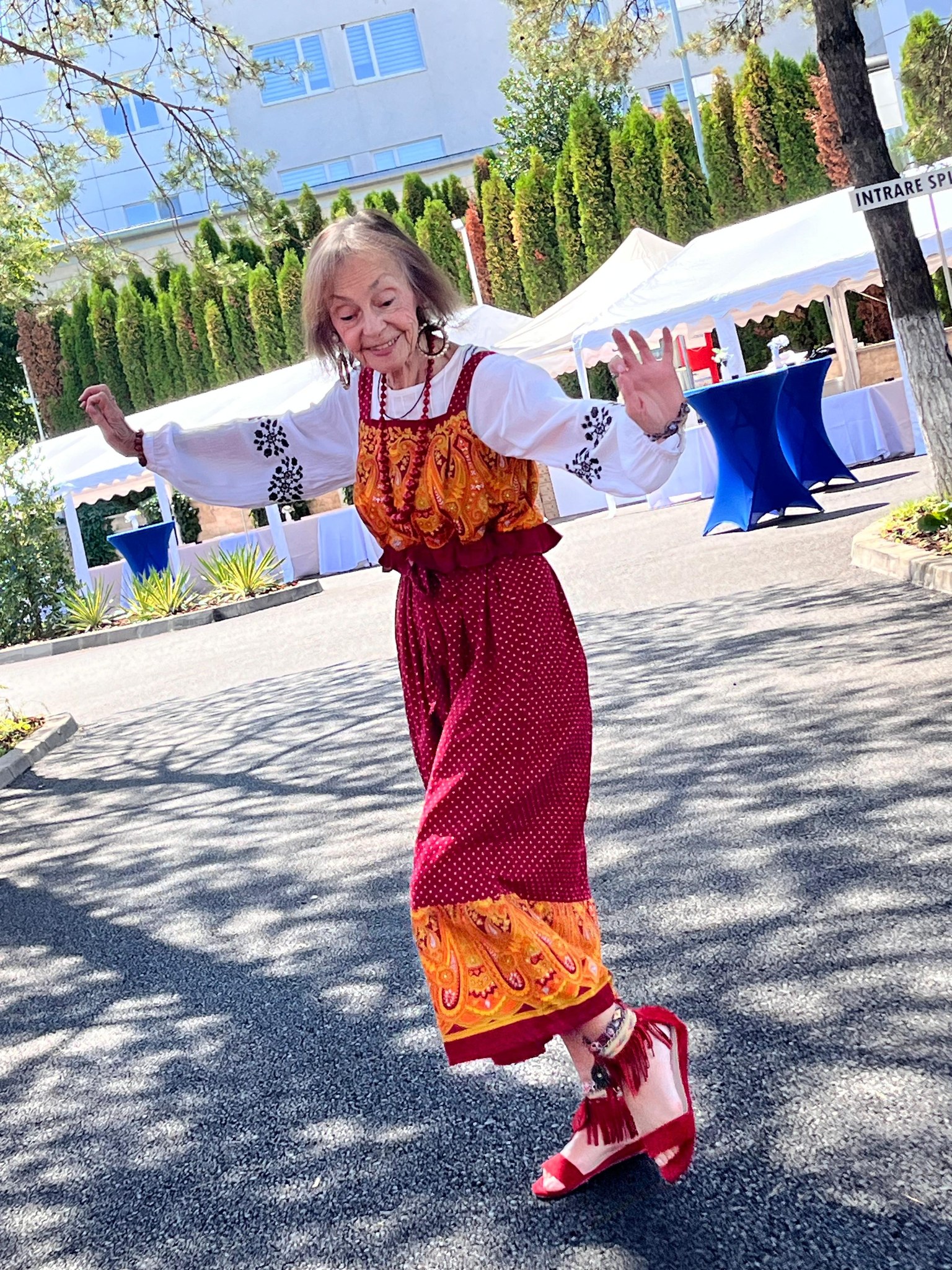Marina Voica veselă, dansând afară, la soare, îmbrăcată în haine fiu colorate și încălțată în sandale roșii, asortate cu ținuta