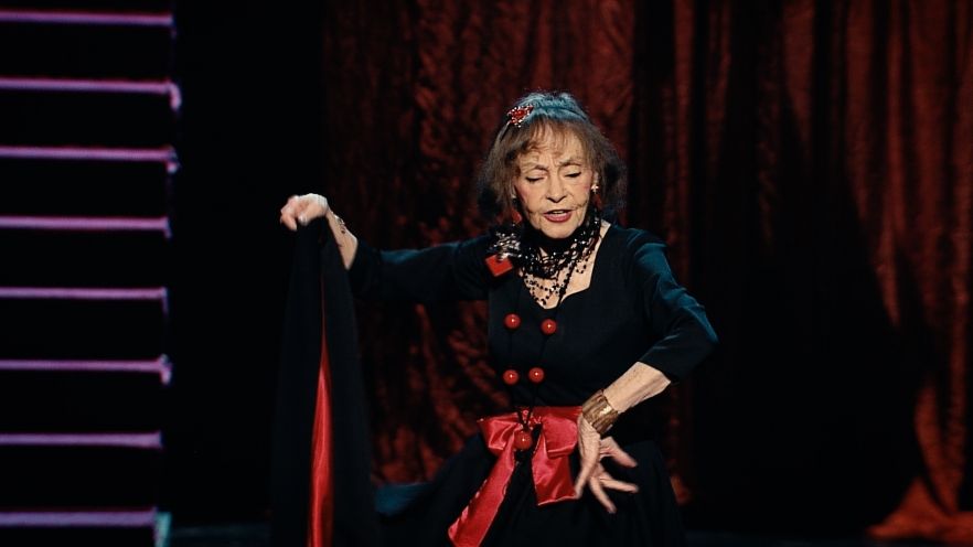 Marina Voica pe scenă, cântând și dansând, îmbrăcată într-o rochie neagră, cu fundă roșie în talie, purtând un colier masiv la gât și o brățară aurie de mari dimensiuni