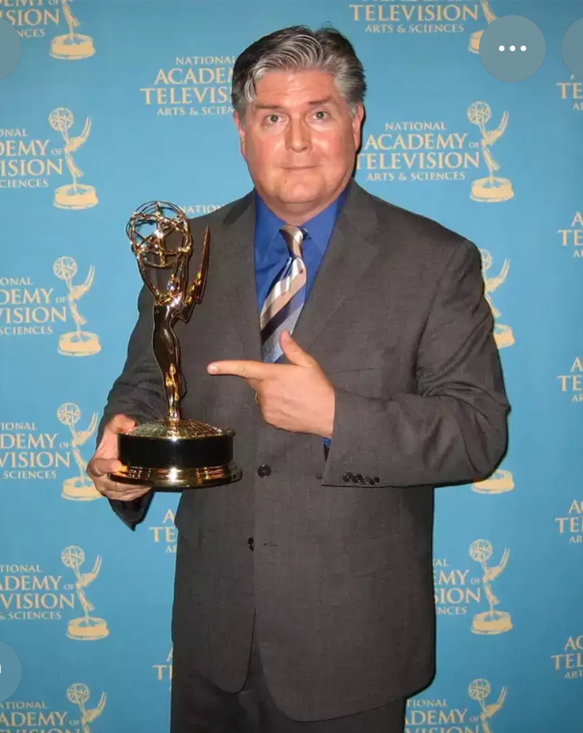 Jim Ward &icirc;ntr-un costum gri, cu cămașă albastră pe dedesubt și cravată &icirc;n dungi oblice colorate, țin&acirc;nd &icirc;n m&acirc;nă un trofeu