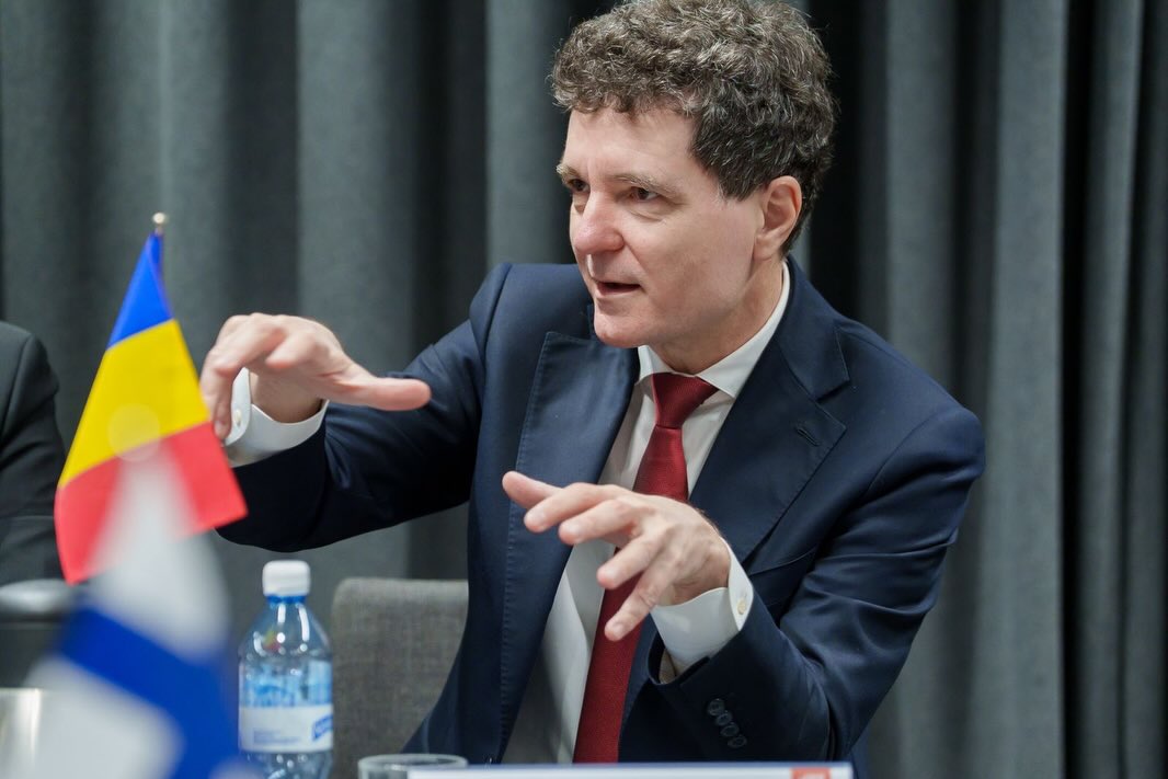 Președintele Nicușor Dan cu steagul Rom&acirc;niei &icirc;n față, gesticul&acirc;nd &icirc;n timp ce ținea un discurs &icirc;n fața unei audiențe, &icirc;mbrăcat &icirc;n costum &icirc;nchis la culoare și cămașă albă, pe care a accesorizat-o cu o cravată vișinie