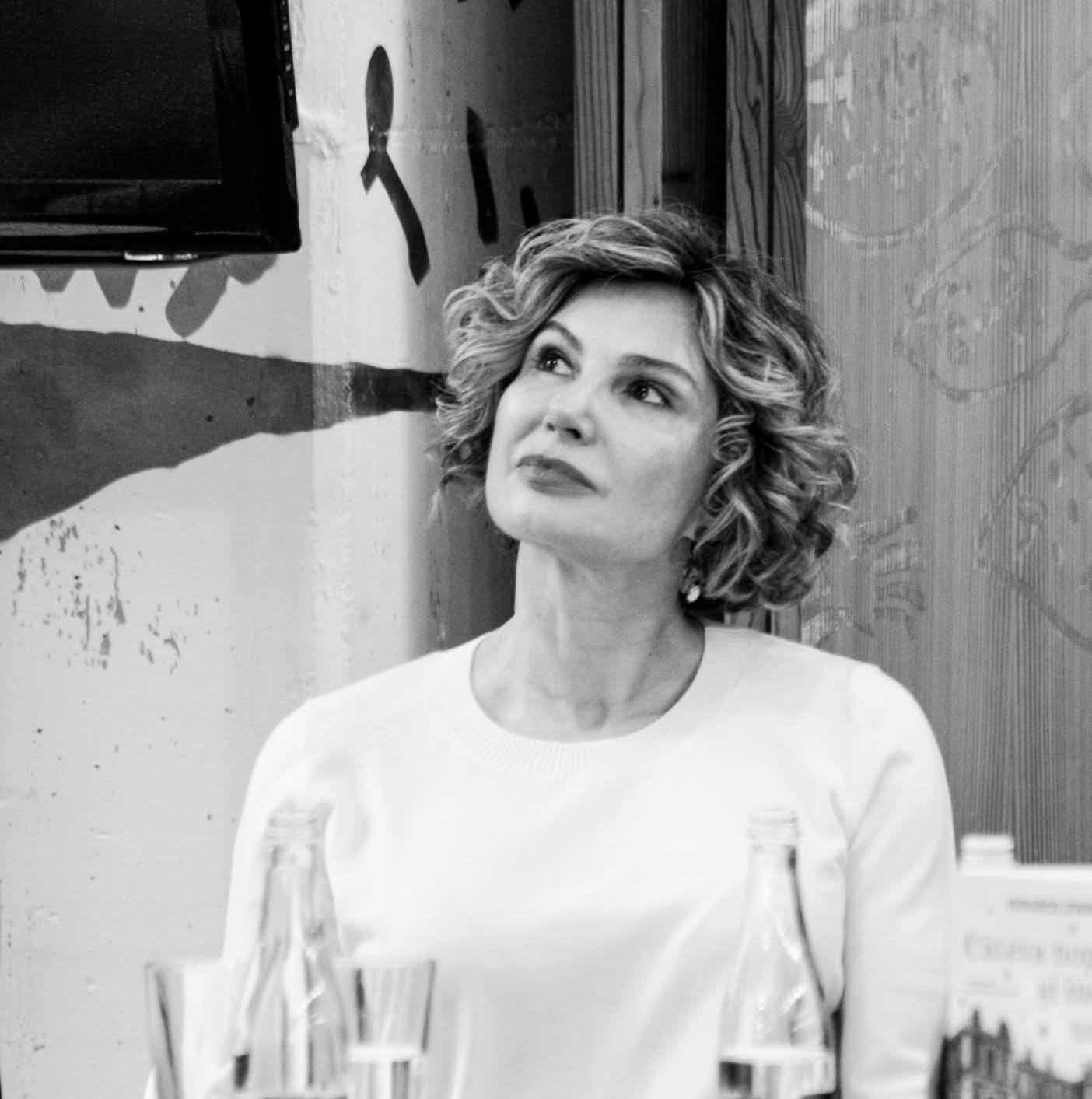 Imagine alb-negru cu Ana Maria Lucescu, soția lui Răzvan Lucescu, &icirc;mbrăcată &icirc;n alb, st&acirc;nd la o masă și privind &icirc;ntr-o parte