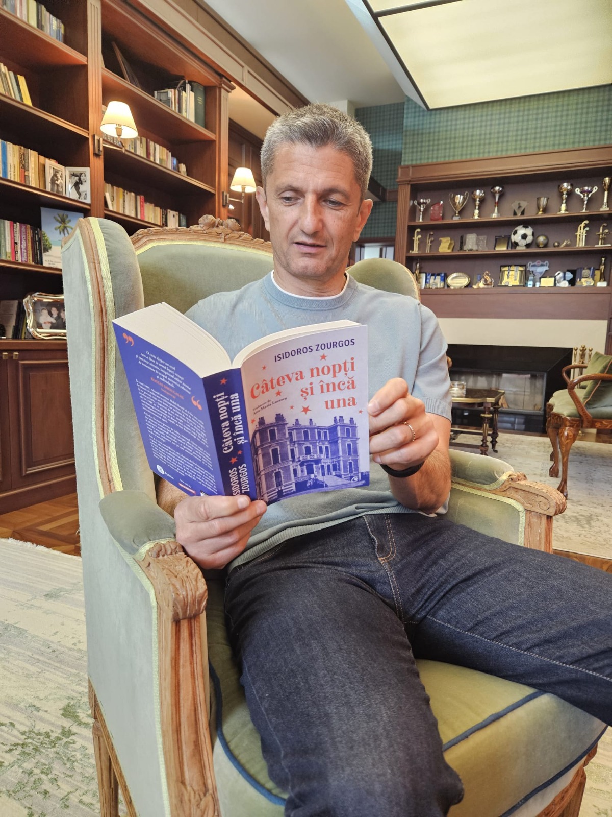 Răzvan Lucescu st&acirc;nd pe un fotoliu, răsfoind cartea &bdquo;C&acirc;teva nopți și &icirc;ncă una&rdquo;, tradusă de soția sa