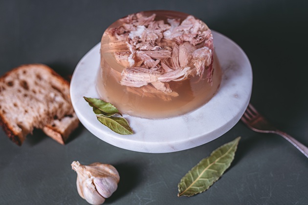 Piftie plină de carne, așezată pe o farfurie albă, rotundă, &icirc;n jurul căreia sunt așezate frunze de dafin, un cățel de usturoi, două felii de p&acirc;ine și o furculiță