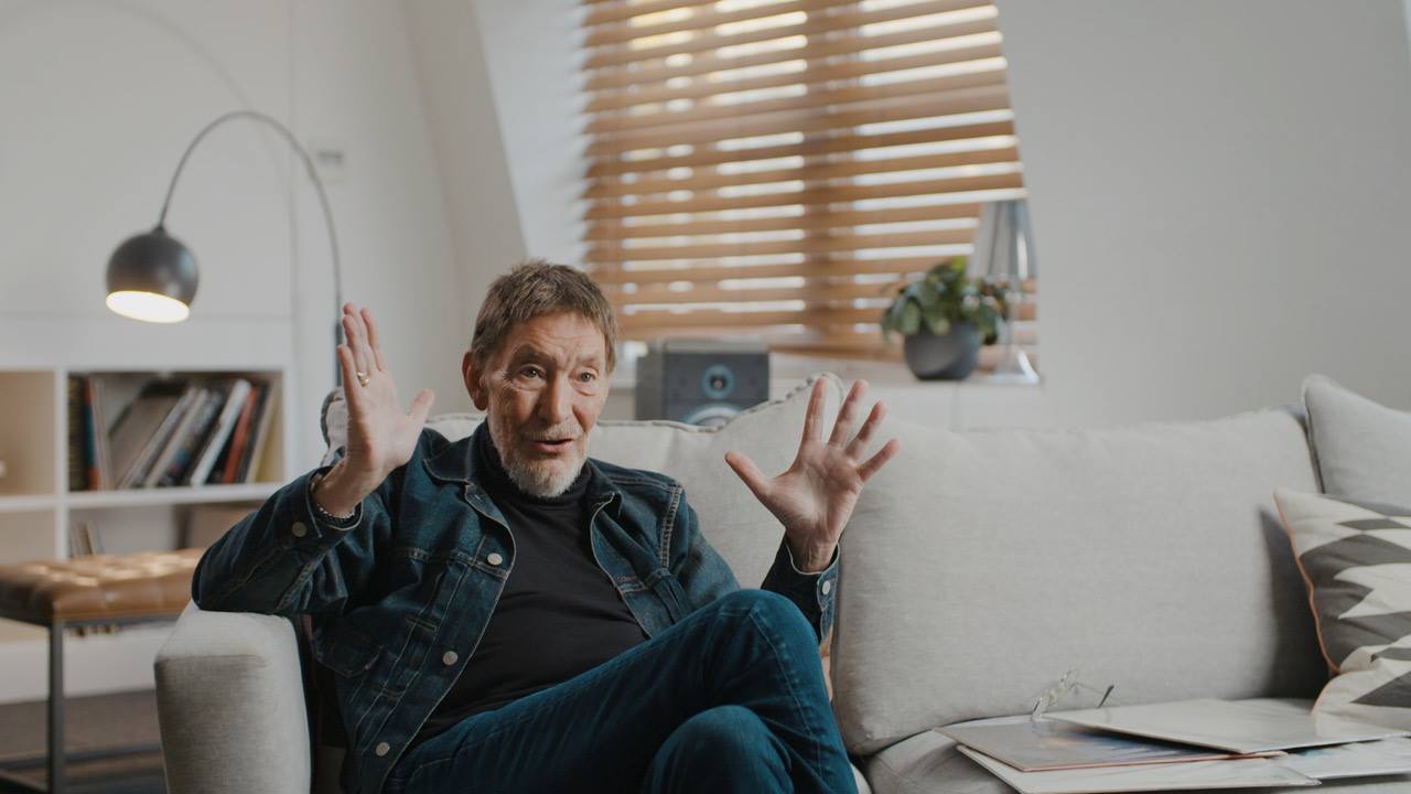 Chris Rea z&acirc;mbitor, &icirc;mbrăcat &icirc;n blugi, tricou negru și geacă de blugi, gesticul&acirc;ng &icirc;n timpul unui interviu, st&acirc;nd relaxat pe o canapea bej