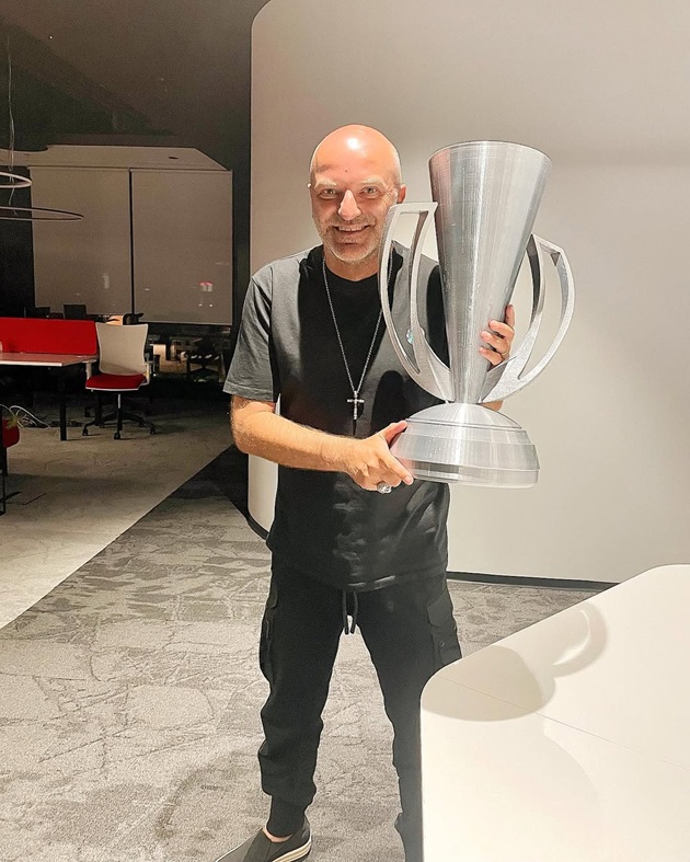 Dan Capatos z&acirc;mbitor, &icirc;mbrăcat &icirc;n negru, cu o cruce la g&acirc;t, țin&acirc;nd &icirc;n m&acirc;nă un trofeu argintiu de mari dimensiuni