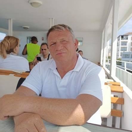 Dănuț Iacob pe o terasă, st&acirc;nd la masă, cu m&acirc;inile &icirc;ncrucișate, &icirc;mbrăcat &icirc;n alb