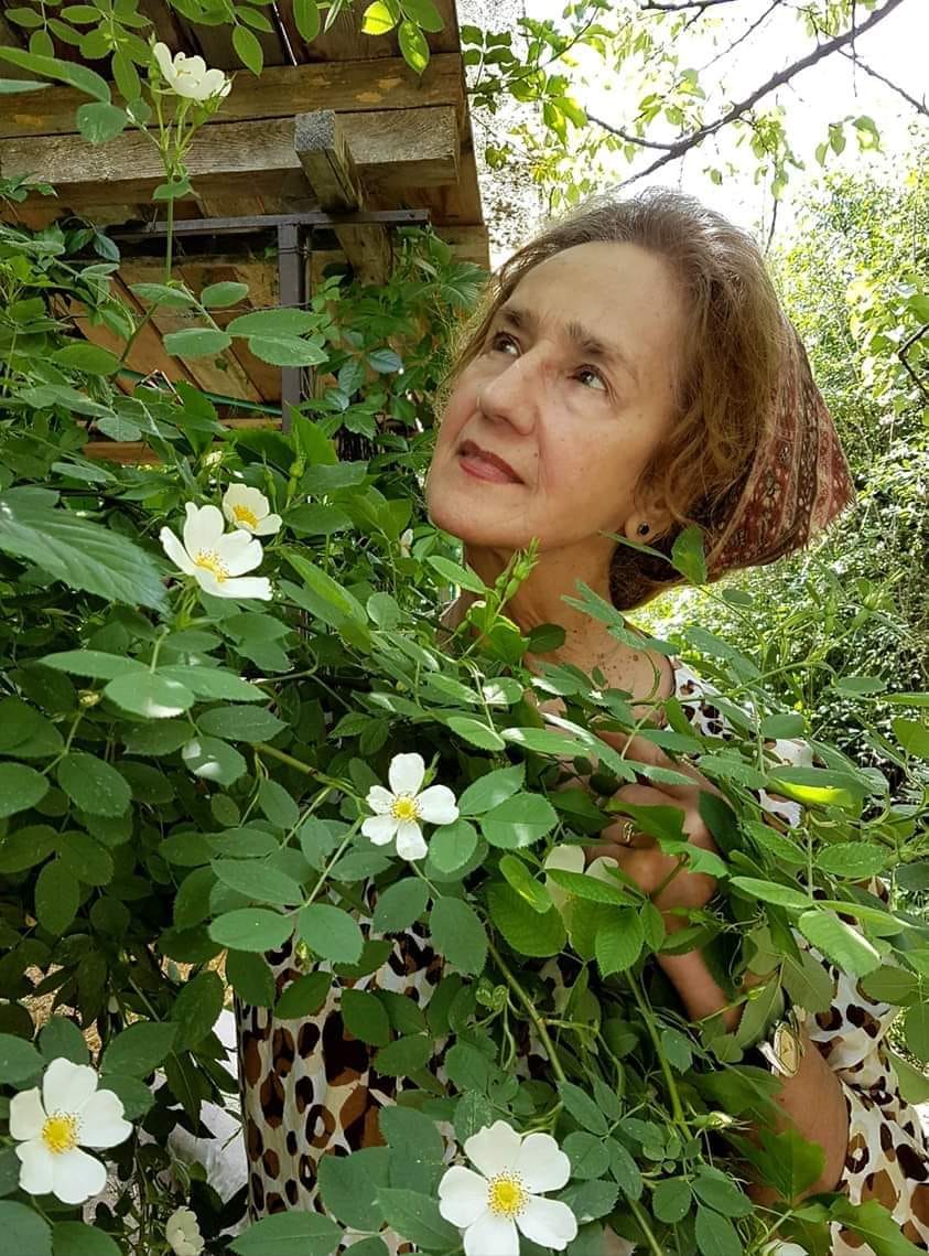 Sofia Vicoveanca admir&acirc;nd un tufiș cu flori, &icirc;ntr-o curte plină de verdeață și de copaci