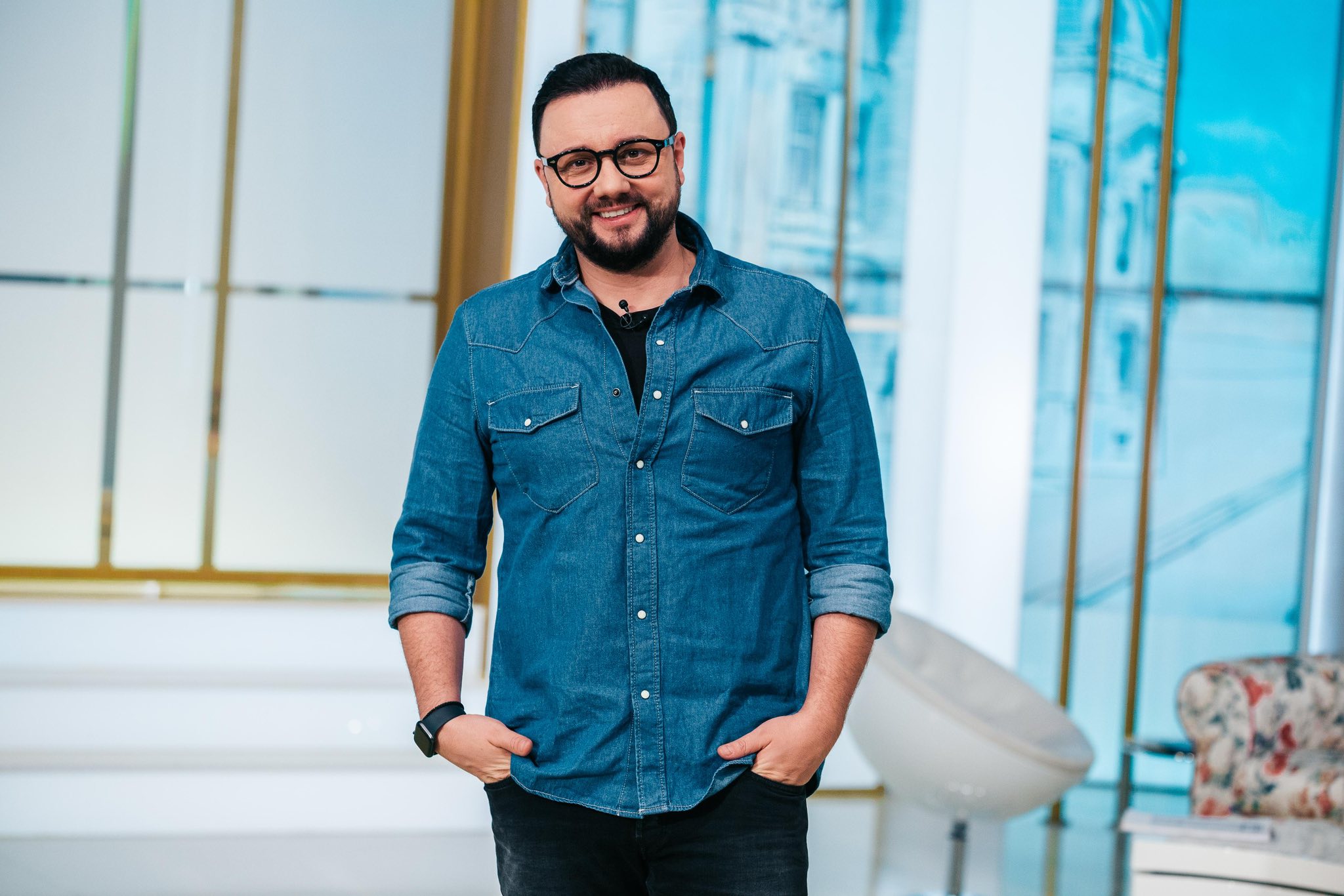 Cătălin Măruță z&acirc;mbitor, st&acirc;nd cu m&acirc;inile &icirc;n buzunar, &icirc;n platoul emisiunii sale de la ProTV, &icirc;mbrăcat &icirc;n pantaloni negri și cămașă albastră din denim