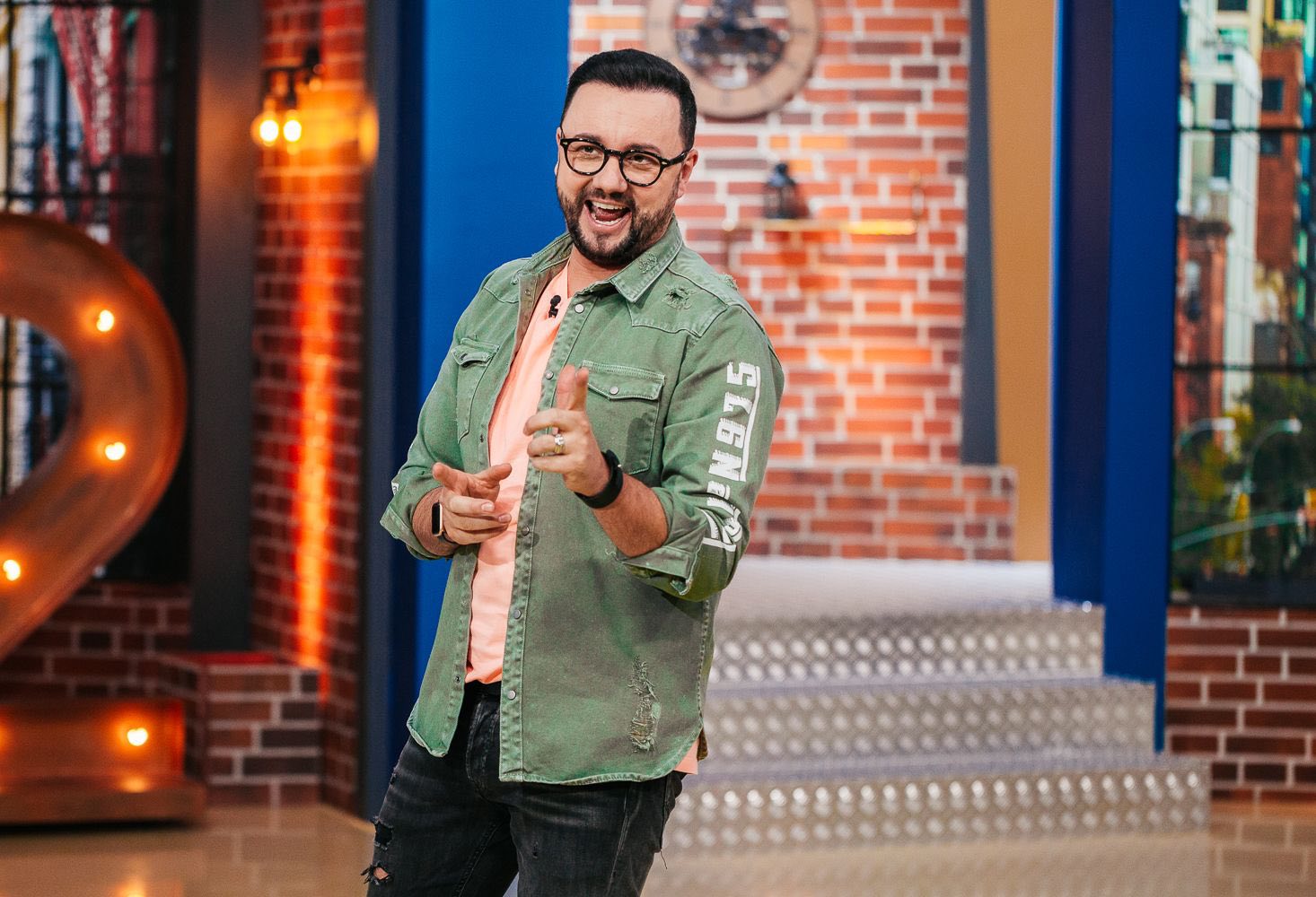 Cătălin Măruță, vesel și z&acirc;mbitor, &icirc;n platoul emisiunii &bdquo;La Măruță&rdquo;, &icirc;mbrăcat &icirc;n pantaloni negri, cămașă verde și tricou portocaliu deschis