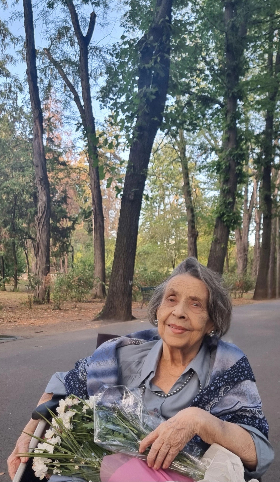Mama Biancăi Brad în parc, la plimbare, în scaun cu rotile, ținând în brațe două buchete de flori albe