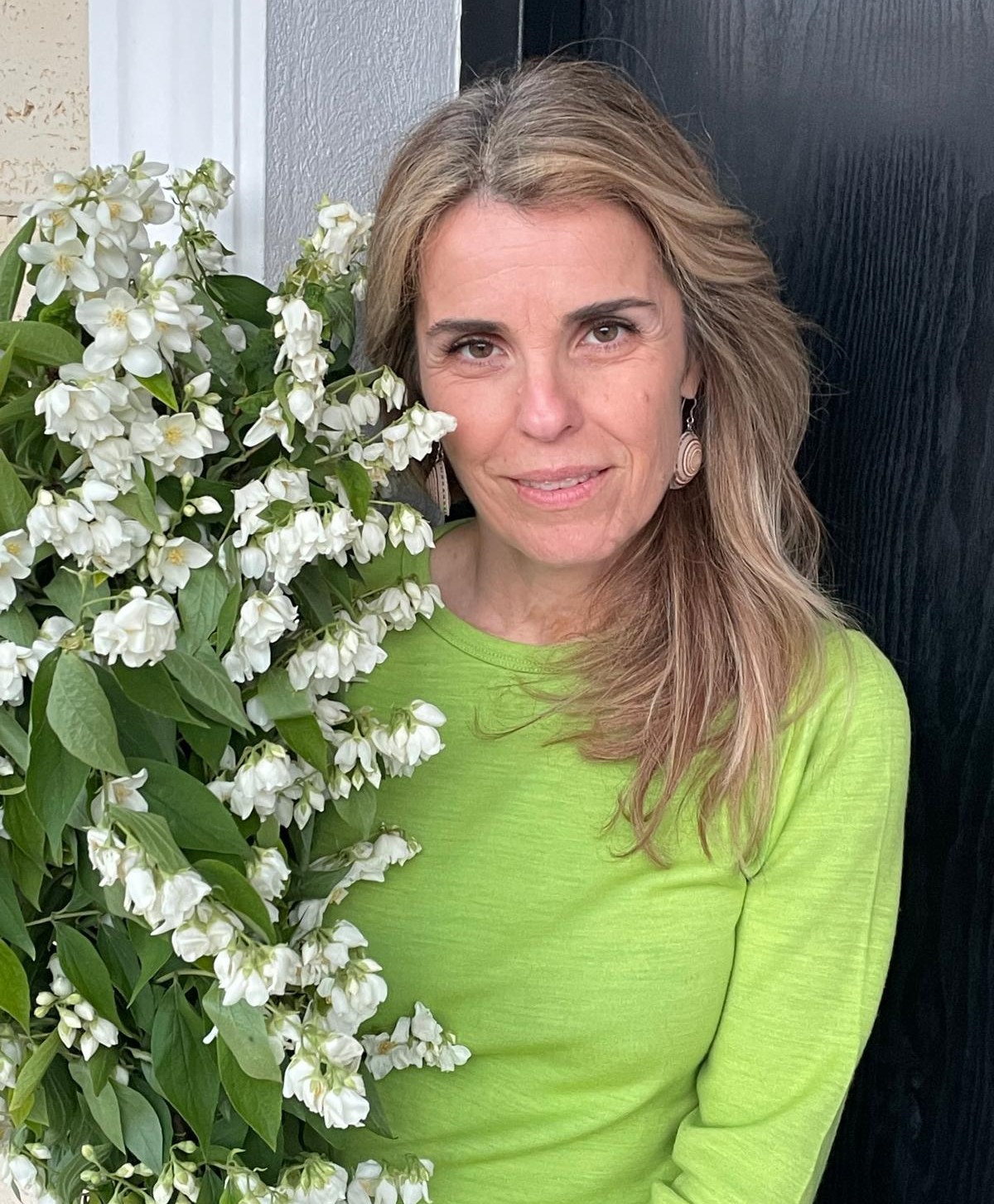 Cristela Georgescu, soția lui Călin Georgescu, &icirc;ntr-o bluză verde lime, st&acirc;nd cu jumătate de corp ascuns după o plantă de mari dimensiuni, cu flori albe și frunze lungi