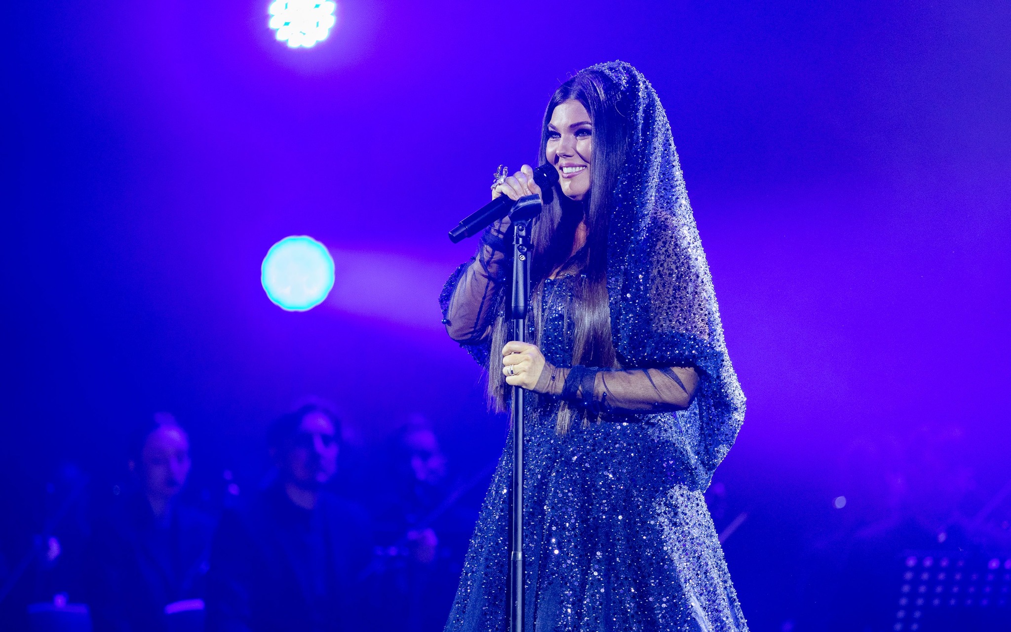 Paula Seling z&acirc;mbitoare pe scenă, cu o eșarfă neagră pe cap, &icirc;mbrăcată &icirc;n rochie cu paiete, țin&acirc;nd str&acirc;ns microfonul