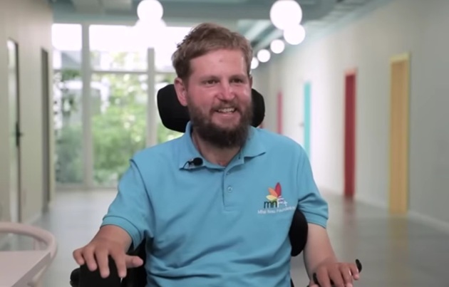 Mihai Neșu z&acirc;mbitor, &icirc;ntr-un tricou bleu, st&acirc;nd &icirc;n scaun cu rotile, &icirc;n centrul pe care l-a construit pentru a ajuta alte persoane cu dizabilități