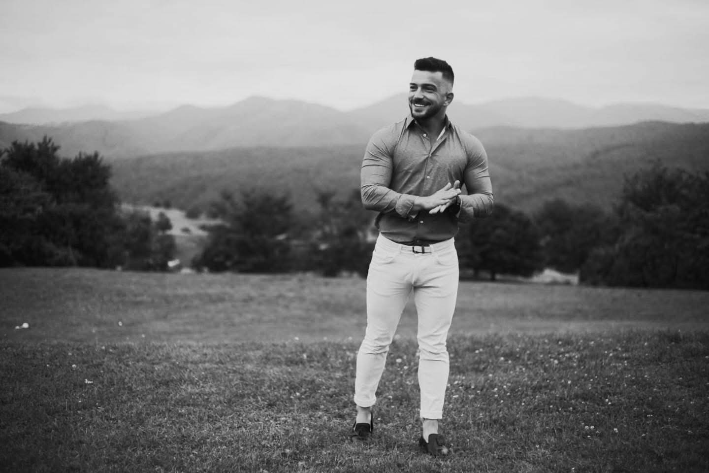 Imagine alb-negru cu Andrei Perneș z&acirc;mbitor, pe o pajiște, &icirc;mbrăcat &icirc;n pantaloni albi și cămașă &icirc;nchisă la culoare, &icirc;n timp ce &icirc;și freca palmele