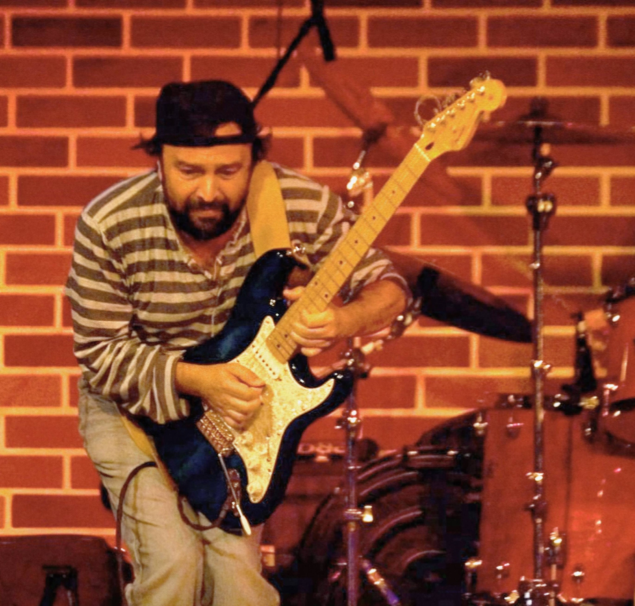 Ioan&nbsp;Gyuri&nbsp;Pascu in bluza cu dungi si cu sapca pe cap canta la chitara cu un perete de caramizi in spate