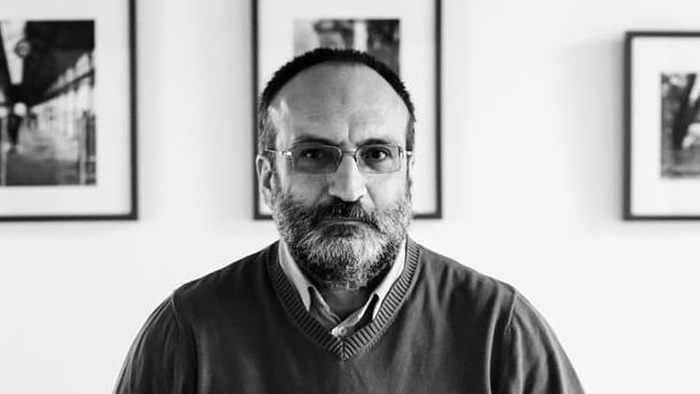 Imagine alb-negru cu jurnalistul Victor Kapra st&acirc;nd serios, &icirc;n fața unui perete alb, cu tablouri de diverse mărimi