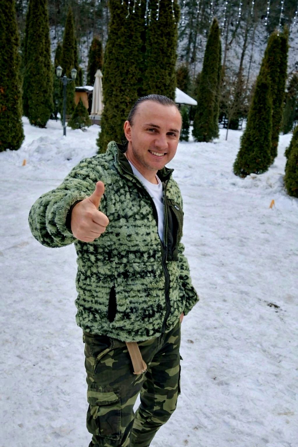Mihai Trăistariu z&acirc;mbitor, &icirc;mbrăcat &icirc;n verde și negru, st&acirc;nd afară, &icirc;nconjurat de zăpadă.