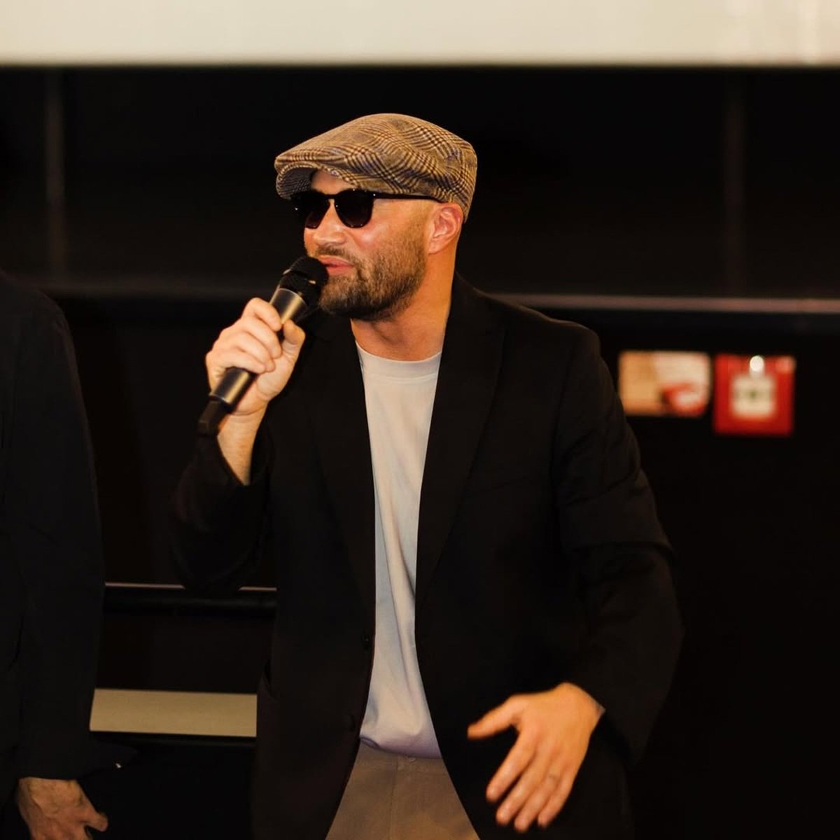 Mihai Bendeac pe scenă, vorbind la microfon &icirc;n fața unui public, cu beretă pe cap și ochelari de soare la ochi