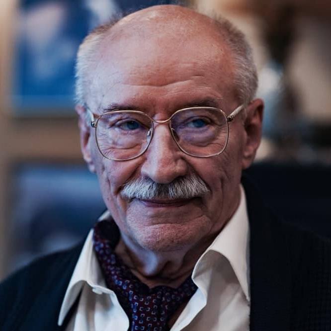 Portret al lui Victor Rebengiuc &icirc;n sacou negru, cămașă albă și cravată cu picățele, purt&acirc;nd ochelari de mari 