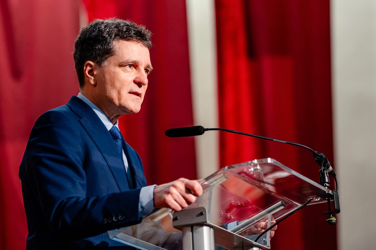 Nicușor Dan fotografiat din profil la un pupitru, vorbind la microfon în fața unei audiențe, în timp ce era îmbrăcat într-un costum albastru, cu cravată asortată