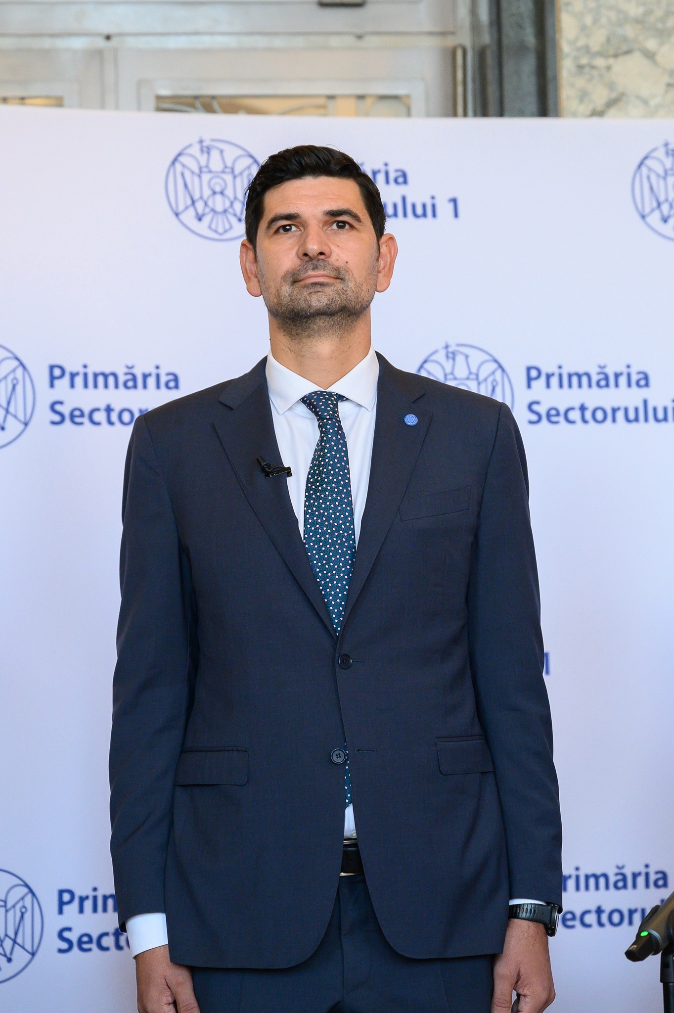 George Tuță, primarul Sectorului 1, &icirc;mbrăcat &icirc;ntr-un costum albastru &icirc;nchis, cu o cravată &icirc;n aceeași culoare cu picățele albe și cămașă albă, st&acirc;nd l&acirc;ngă un panou pe care scrie Primăria Sectorului 1