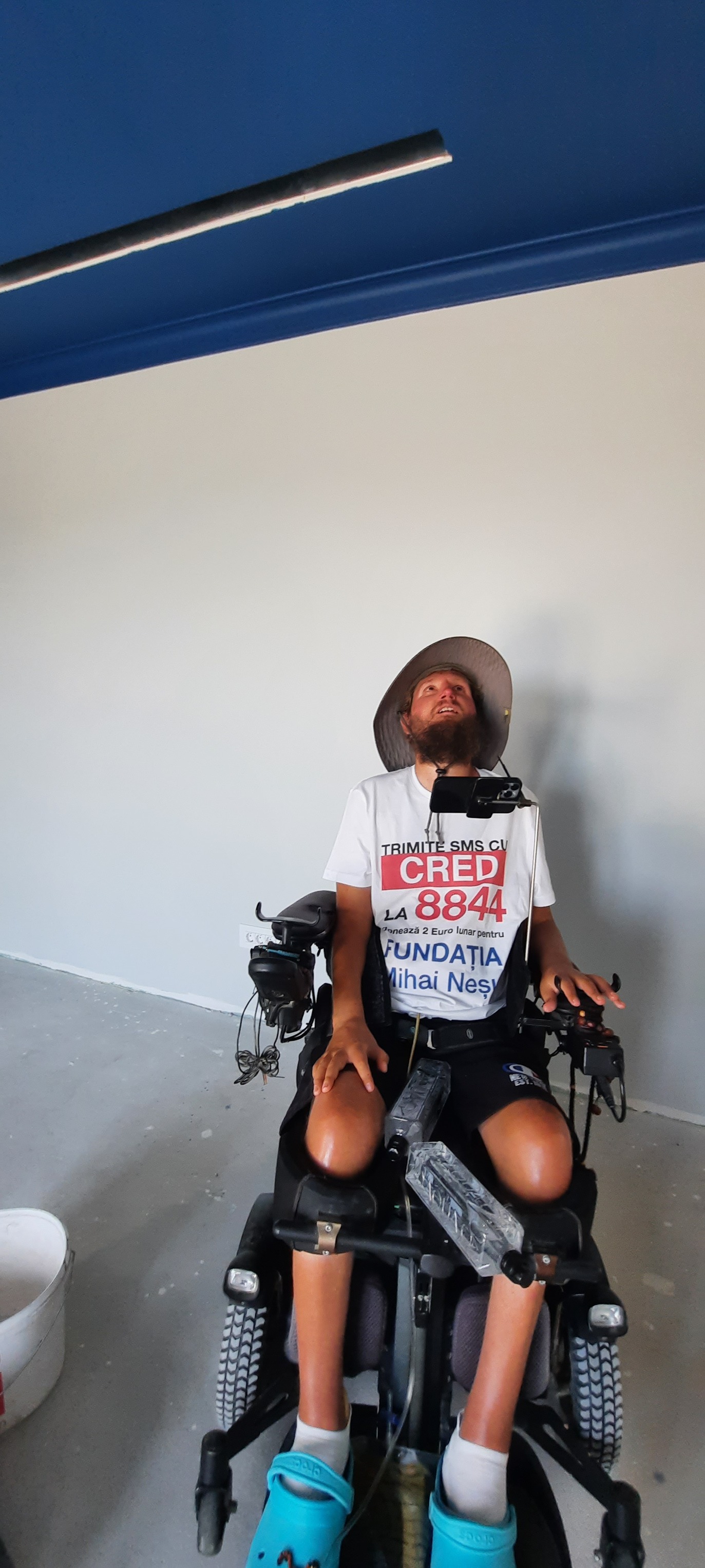 Mihai Nesu în scaun cu rotile, în pantaloni scurți negri, purtând în partea de sus un tricou cu numărul la care oamenii pot trimite SMS pentru a dona fundației sale