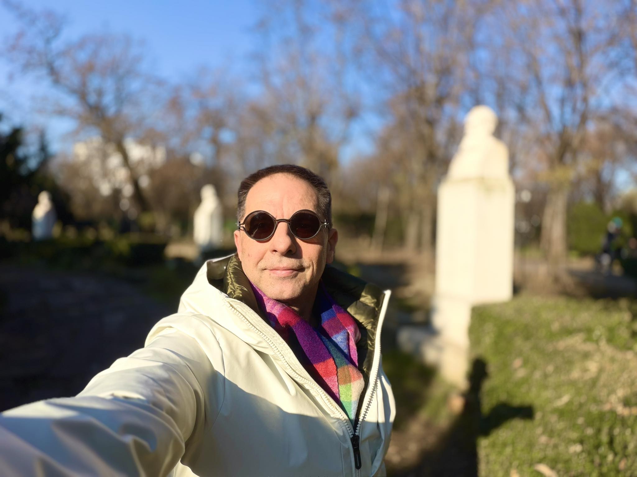 Dan Negru făc&acirc;ndu-și un selfie cu ochelarii de soare la ochi, &icirc;n geacă albă și cu fular colorat, st&acirc;nd la soare &icirc;ntr-un parc, &icirc;n timp ce era &icirc;nconjurat de mai multe statui