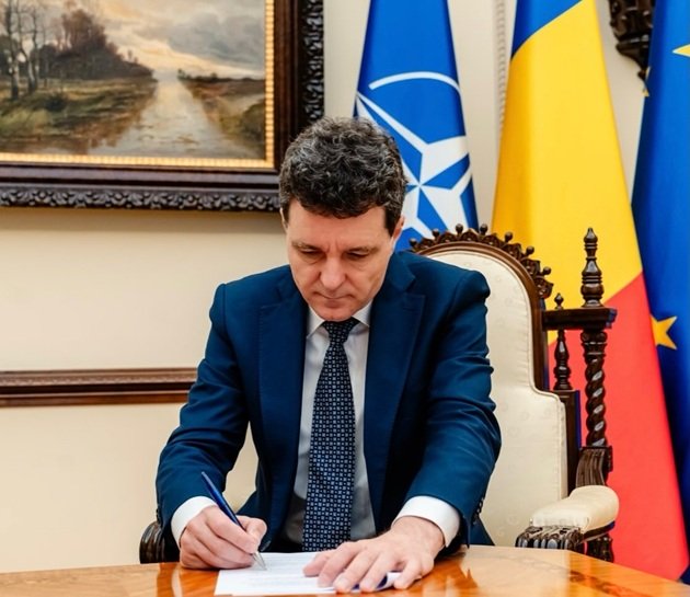 Nicușor Dan, președintele Rom&acirc;niei, st&acirc;nd la birou și semn&acirc;nd un act oficial, av&acirc;nd &icirc;n spate drapelul Rom&acirc;niei, drapelul NATO și drapelul UE